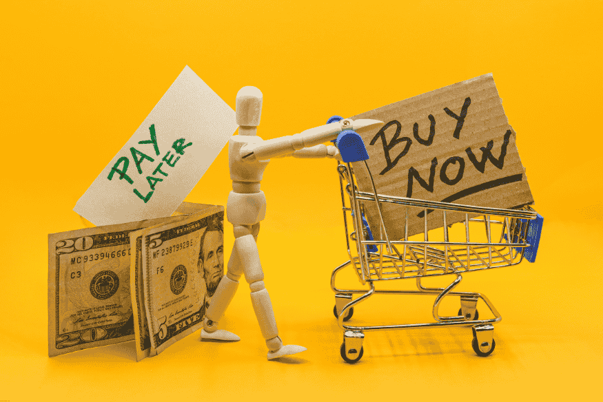 Wooden mannequin pushing a shopping cart with “Buy Now” and “Pay Later” signs beside dollar bills