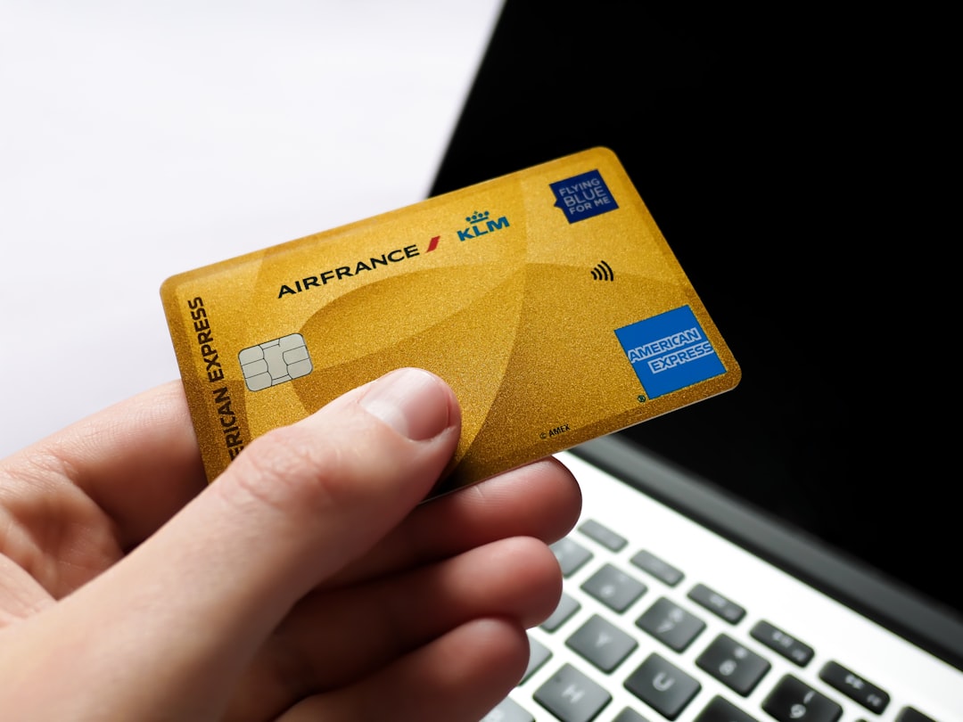 Hand holding a gold american express card near laptop.