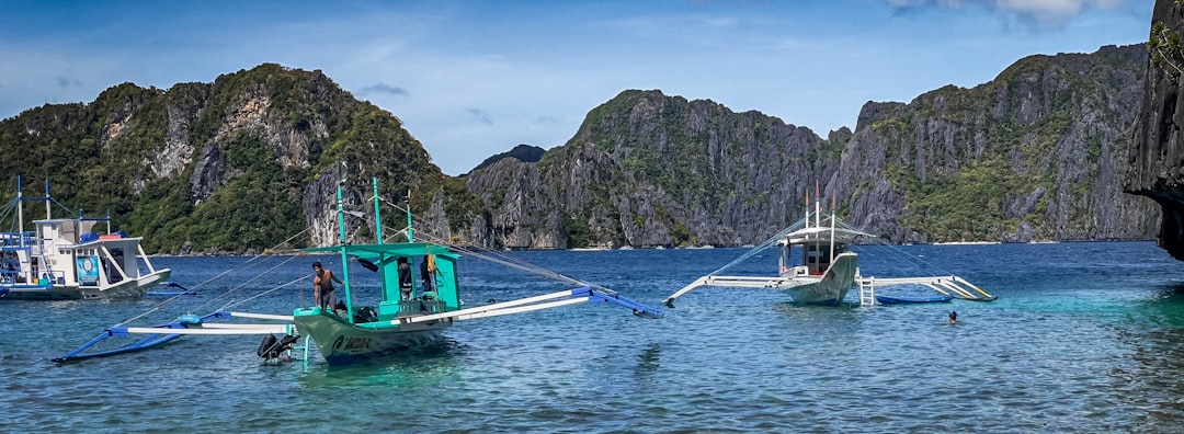 Boats bob on blue water near towering cliffs.