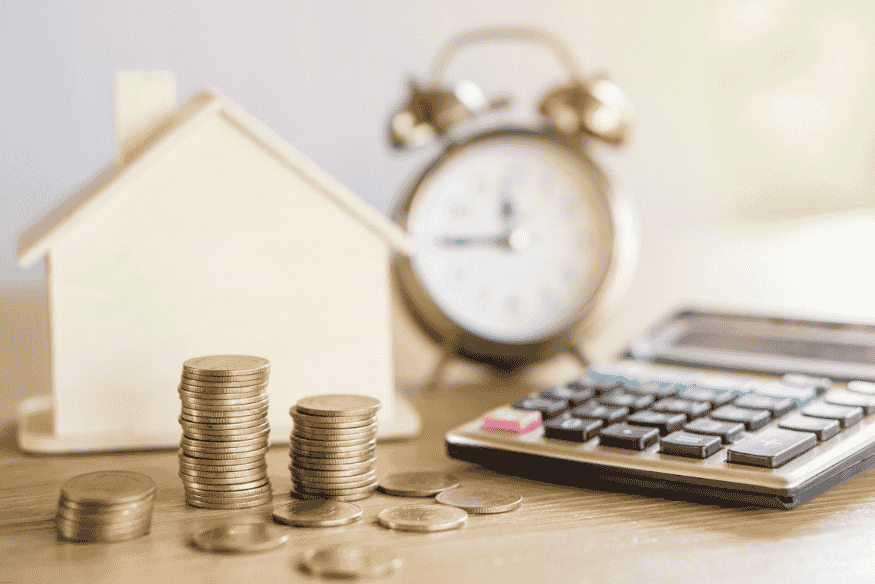 Stacked coins, calculator, small house model, and clock representing time-based financial growth.