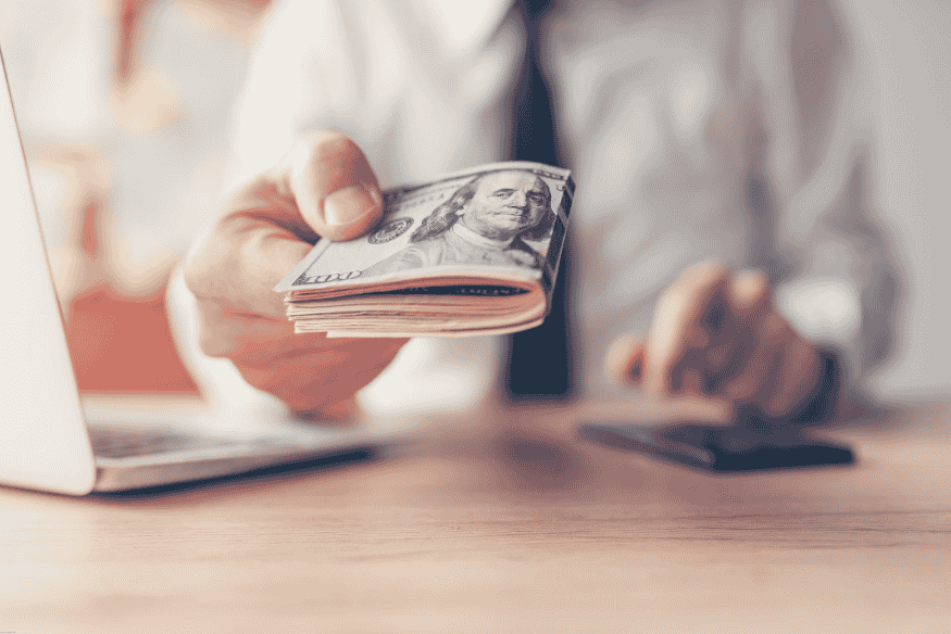 Businessman handing cash in US dollars across a desk, with a laptop in the background.