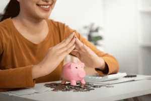 Woman smiling while protecting a piggy bank with her hands over coins on a desk.
